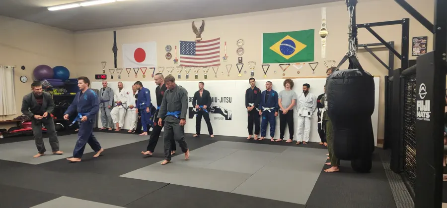 Adult Brazilian Jiu-Jitsu class in gis lined up and drilling on the mats, with the heavy bag and cage in the background